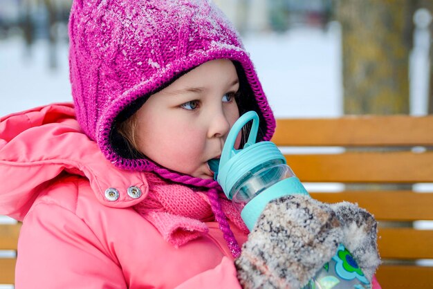 Un bambino nel parco in inverno beve il tè giocando sulla neve una bambina bianca di cinque anni beve una calda te...