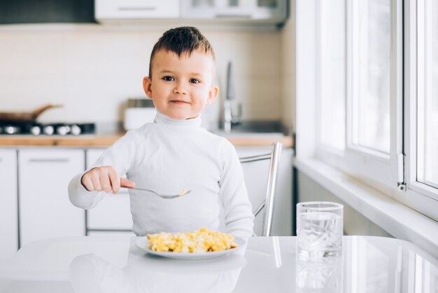 Un bambino nel pomeriggio in una cucina a luce bianca con un maglione bianco mangia una frittata