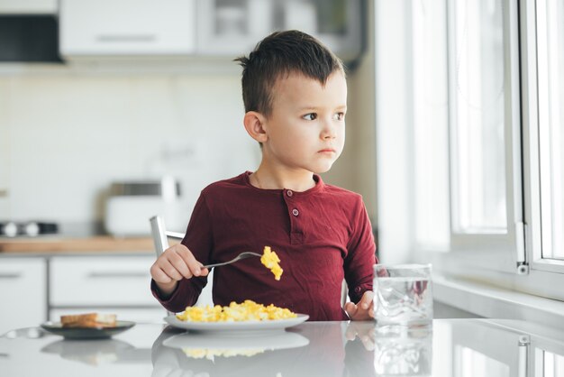 Un bambino nel pomeriggio in una cucina a luce bianca con un maglione bordeaux mangia una frittata