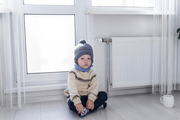 Un bambino piccolo con un maglione e un cappello grigio è inginocchiato vicino a una stufa con un termostato