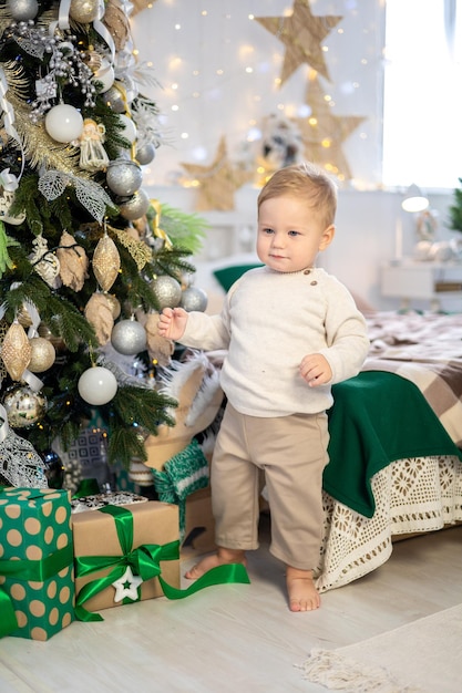 Un bambino piccolo con un maglione lavorato a maglia si trova vicino al letto sullo sfondo di un albero di Natale decorato a festa a casa il bambino festeggia il Natale e il nuovo anno a casa