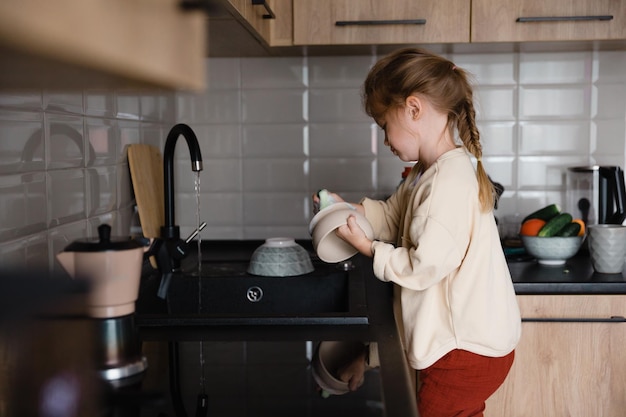 Un bambino piccolo con uno straccio in mano sta su una sedia vicino al lavandino e lava i piatti Una bambina aiuta sua madre a cucinare e pulire in cucina