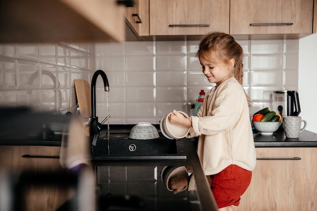 Un bambino piccolo con uno straccio in mano sta su una sedia vicino al lavandino e lava i piatti Una bambina aiuta sua madre a cucinare e pulire in cucina