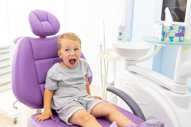 Un bambino piccolo di due anni seduto su una poltrona odontoiatrica lilla con la bocca aperta.