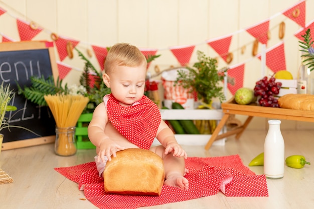 Un bambino piccolo di sei mesi è seduto sul pavimento della cucina con una pagnotta di pane