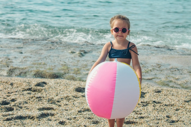 Un bambino piccolo in occhiali da sole gioca con una palla gonfiabile sulla sabbia. ragazza sulla spiaggia. Vacanze estive al mare. Viaggia in paesi caldi