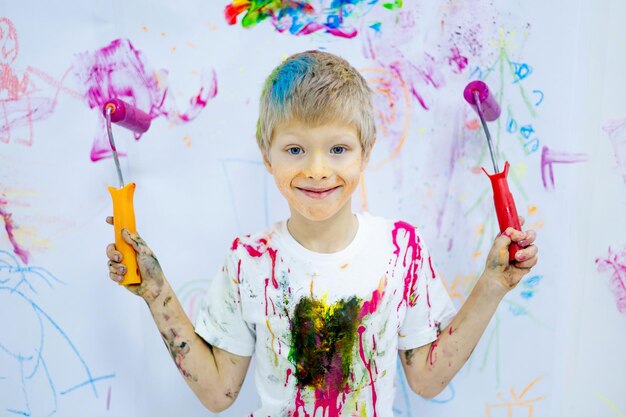 Un bambino piccolo macchiato e dipinto con vernice tiene un pennello e un rullo per disegnare