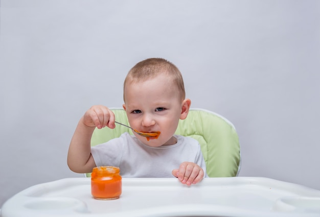 Un bambino piccolo mangia purè di carote a un tavolo e guarda la telecamera su un bianco isolato