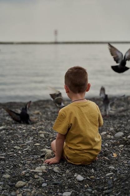 Un bambino piccolo si siede vicino al mare e dà da mangiare ai piccioni.