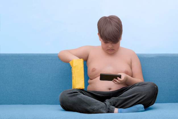 Un bambino pieno si siede su un divano a suonare un telefono e mangiare snack. Obesità nei bambini. Copia spazio.