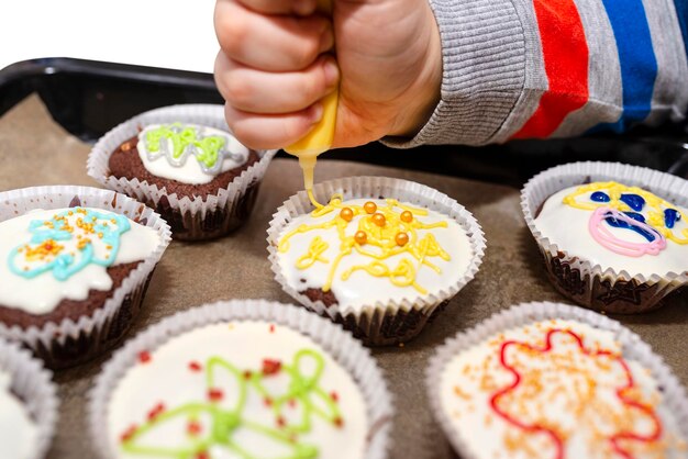 Un bambino schiaccia la glassa colorata da un tubo su cupcakes marrone cioccolato coperti di glassa bianca