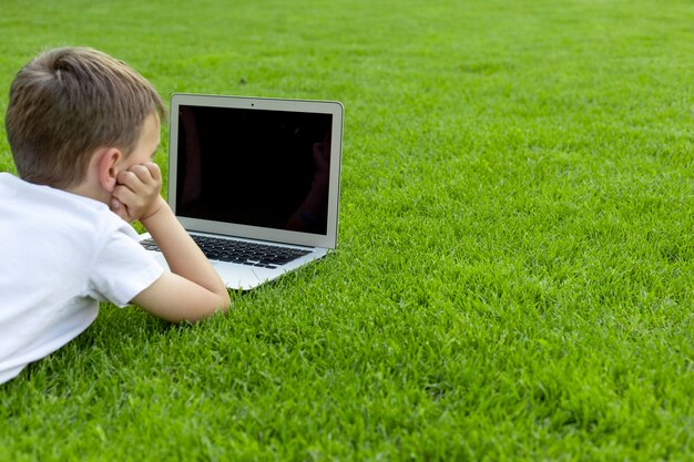 Un bambino si siede sull'erba e studia con un laptop online, godendosi la natura