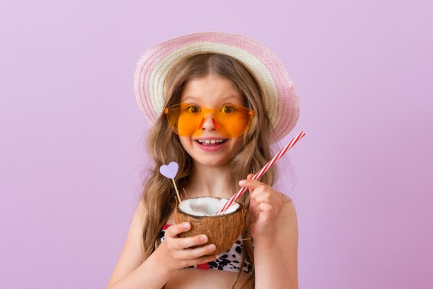 Un bambino sorridente con cappello e occhiali beve un cocktail di cocco. Muro isolato rosa.