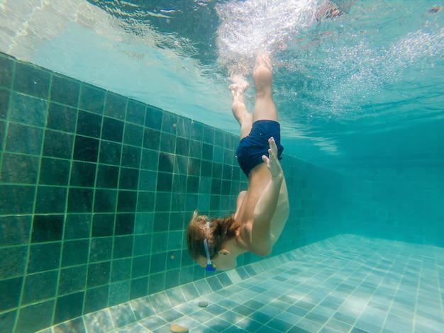 Un bambino sta nuotando sott'acqua in una piscina, sorride e trattiene il respiro, con gli occhiali da nuoto