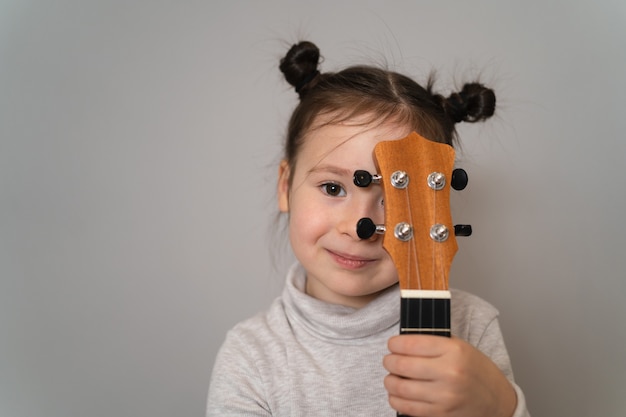 Un bambino tiene in mano un ukulele. Piccoli bambini creativi. La ragazza impara a suonare uno strumento online. Bambino creativo che tiene una chitarra vicino al suo viso