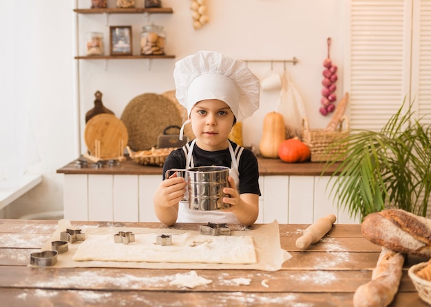 Un bambino vestito da chef setaccia la farina al setaccio in una cucina di legno