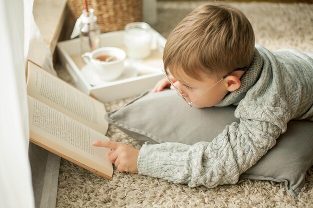 Un bel bambino con un maglione lavorato a maglia sta leggendo vicino alla finestra con una tazza di tè caldo. Accogliente. Autunno. Umore autunnale.