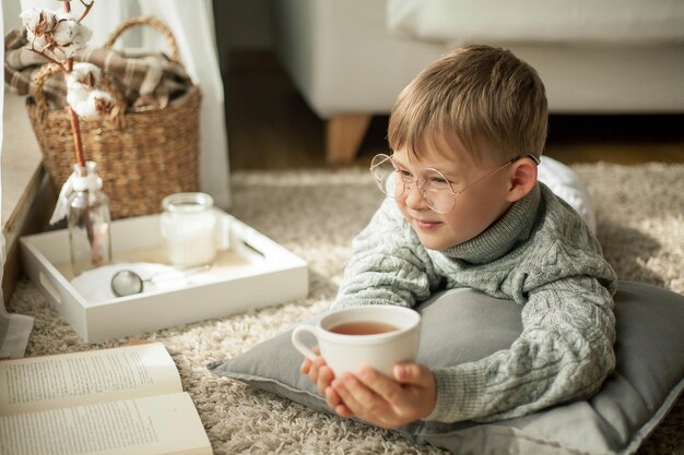 Un bel bambino con un maglione lavorato a maglia sta leggendo vicino alla finestra con una tazza di tè caldo. Accogliente. Autunno. Umore autunnale.