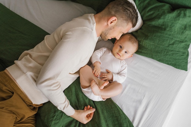 Un bel giovane papà giace a letto in camera da letto e abbraccia il suo amorevole ragazzo di sei mesi che ride Il concetto di amore incondizionato e paternità