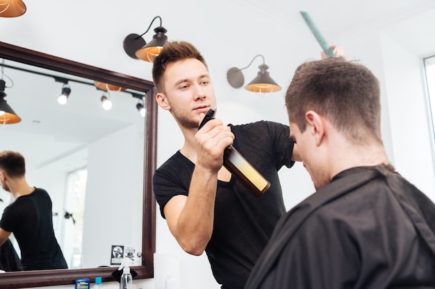 Un bel giovane parrucchiere concentrato spruzza con i capelli d'acqua di un cliente attraente al barbiere