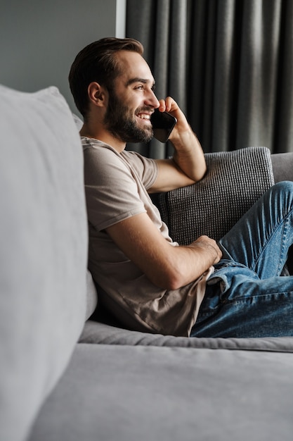 un bel giovane sorridente felice al chiuso a casa sul divano parlando al telefono cellulare.