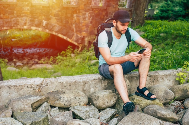 Un bel giovane turista con uno zaino che si riposa durante un viaggio di escursione seduto su una roccia che consulta il suo smartphone