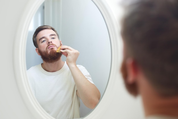 Un bel giovane uomo barbuto europeo uomo brutale con i capelli della barba sta pettinando la barba sul viso con un