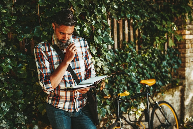 Un bel giovane va in città con la sua bicicletta, in piedi accanto ad essa, in attesa di qualcuno e leggendo un libro.