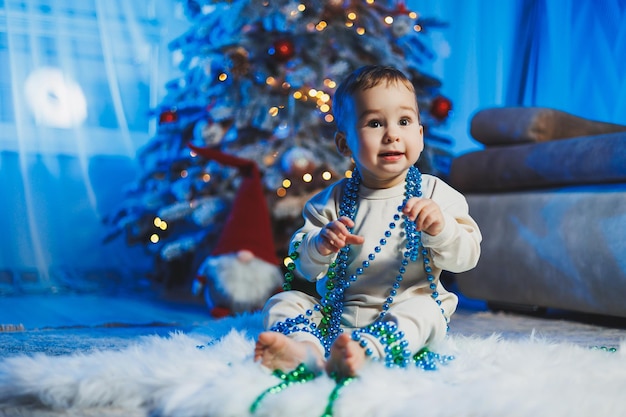 Un bel ragazzino di un anno vestito con un abito beige sullo sfondo di un albero di Natale in un ambiente domestico Le emozioni dei bambini prima di celebrare le vacanze di Capodanno
