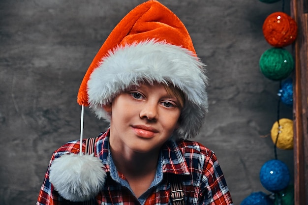 Un bel ragazzo adolescente con il cappello di Babbo Natale che indossa una camicia a scacchi con bretelle. Isolato su uno sfondo scuro con texture.