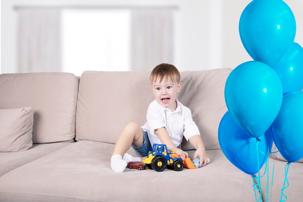 Un bel ragazzo con le emozioni sul viso gioca con i giocattoli su un grande divano vicino alla finestra e palloncini blu pieni di elio Buon compleanno