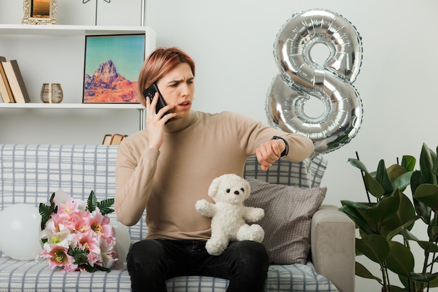 Un bel ragazzo confuso durante la giornata delle donne felici che tiene l'orsacchiotto parla al telefono seduto sul divano nel soggiorno