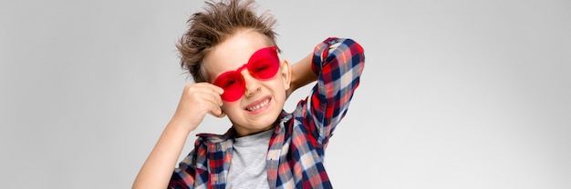 Un bel ragazzo in una camicia a quadri, camicia grigia e jeans si erge su uno sfondo grigio. Un ragazzo in occhiali da sole rossi. Il ragazzo tiene gli occhiali