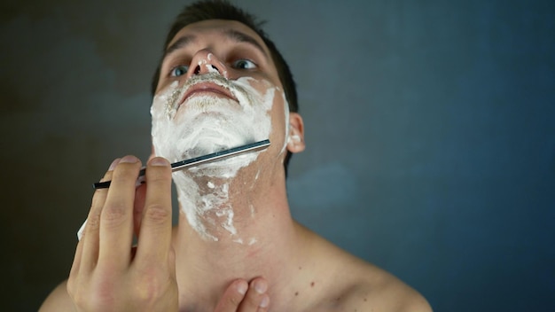 Un bel ragazzo si rade la barba con un rasoio pericoloso Tagliando la barba con una lama vecchio stile in bagno Un bell'uomo macho si è rasato la barba a casa Colpo del primo piano