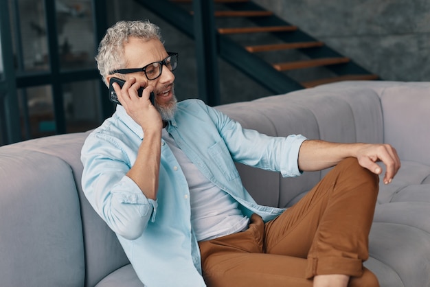 Un bell'uomo anziano in abbigliamento casual e occhiali che parla allo smartphone mentre è seduto sul divano di casa