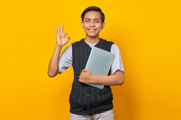 Un bell'uomo asiatico che tiene in mano un laptop e mostra gesticolando un cartello ok su sfondo giallo
