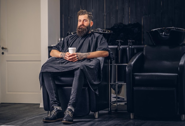 Un bell'uomo barbuto con un tatuaggio sulle braccia beve il caffè prima di lavarsi i capelli in un salone di parrucchiere.