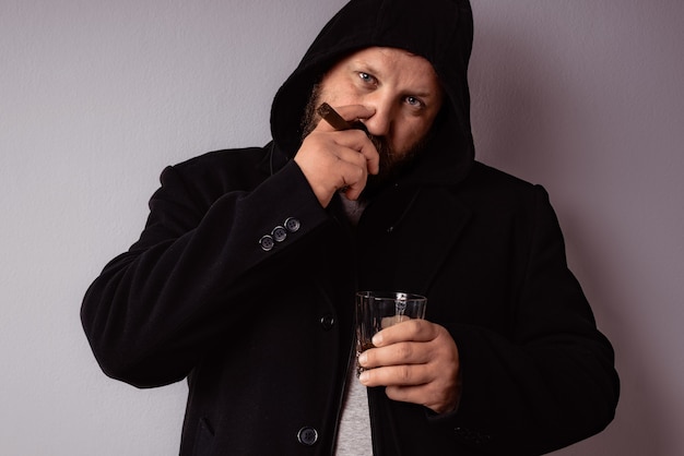 Un bell'uomo barbuto elegante che indossa un cappotto nero beve un liquore forte e fuma il suo sigaro