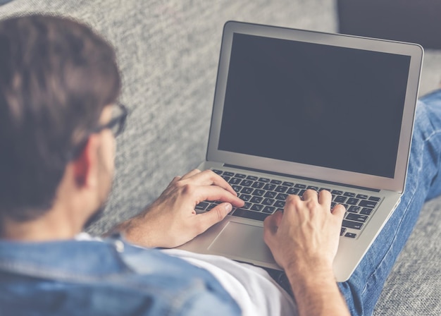 Un bell'uomo con gli occhiali sta usando un laptop mentre è sdraiato sul divano a casa