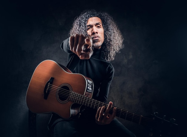 Un bell'uomo con i capelli lunghi tiene in mano la chitarra acustica mentre punta il dito verso il fotografo.