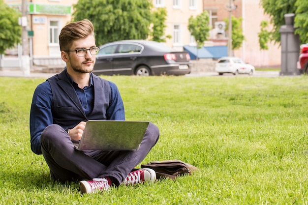 Un bell'uomo con la barba è seduto su un prato e lavora su un laptop