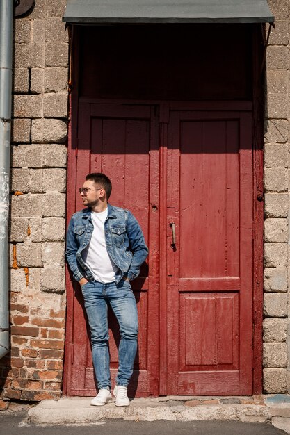 Un bell'uomo con la barba in giacca di jeans e pantaloni di jeans si trova vicino a porte vintage rosse. Ragazzo cresciuto in stile denim