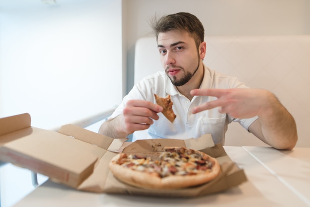 Un bell'uomo con un pezzo di pizza in mano pone sulla macchina fotografica. Uomo seduto a un tavolo vicino alla scatola della pizza