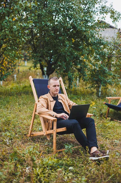 Un bell'uomo con una camicia marrone siede su una sedia pieghevole in legno in giardino e lavora con un computer portatile Mobili ecologici in legno Lavoro a distanza
