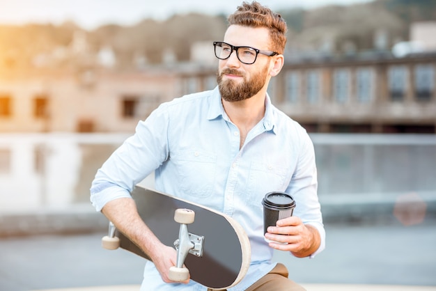 Un bell'uomo d'affari che fa una pausa caffè seduto con lo skateboard sul tetto sullo sfondo industriale