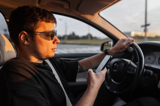 Un bell'uomo d'affari con occhiali da sole in maglietta nera sta usando uno smartphone e guida un'auto al tramonto