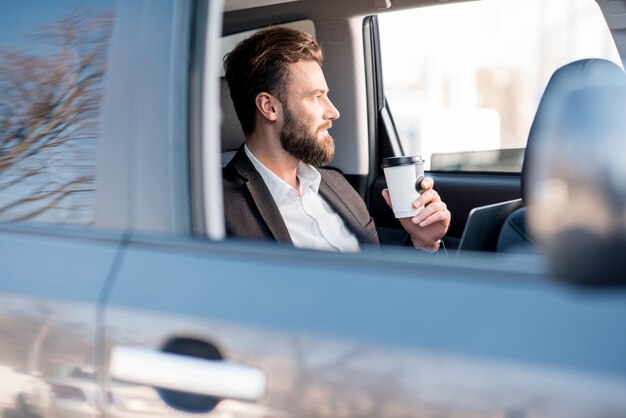 Un bell'uomo d'affari seduto con un caffè per andare sul sedile posteriore dell'auto