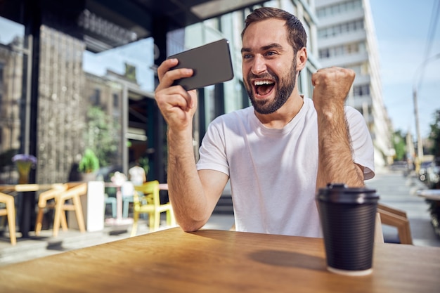 Un bell'uomo è felice di guardare lo smartphone seduto al tavolo con un caffè al bar