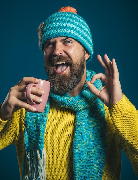 Un bell'uomo hipster con barba in sciarpa cappello e maglione tiene una tazza con caffè caldo o spettacoli di tè