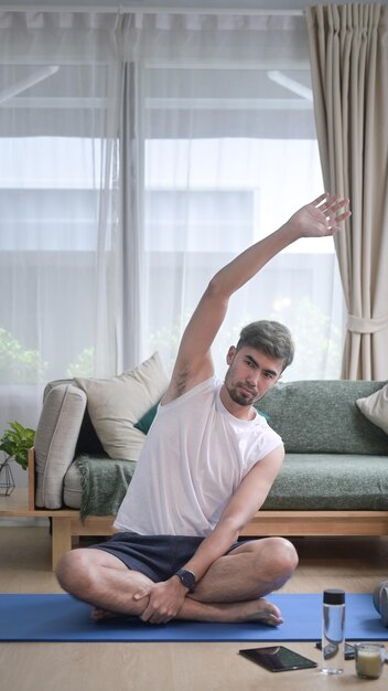 Un bell'uomo in abiti sportivi si sta riscaldando mentre si siede sul tappetino da yoga a casa